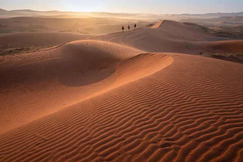 Rippled red sand dunes of Mui Ne at sunrise with distant figures walking in soft golden light